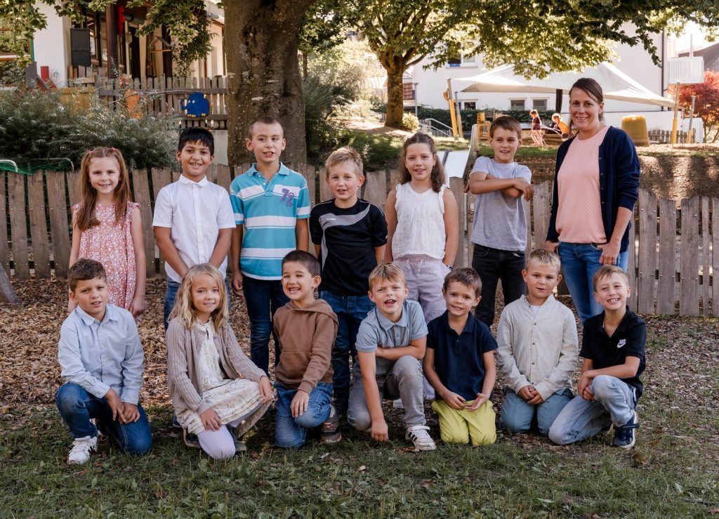 Klassenfoto der Klasse 2 (Tigerklasse) mit Klassenlehrerin Frau Kerstin Rasch vor einem Holzzaun auf dem Schulhof, vorne knien sieben Kinder, dahinter stehen sieben Kinder